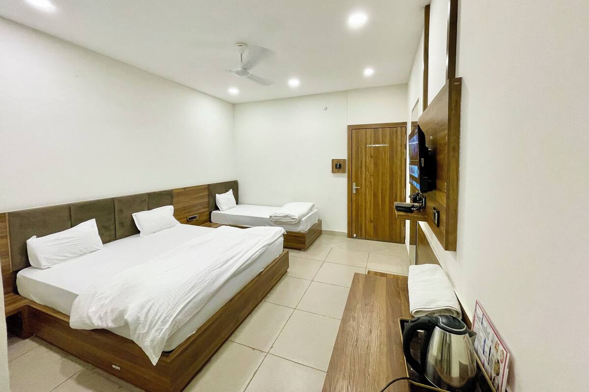 A hotel room with two beds, a wooden headboard, a ceiling fan, a TV mounted on the wall, and a kettle on the side table.