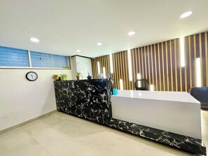 Zona de recepción con mostrador de mármol, un reloj y persianas verticales en la pared.