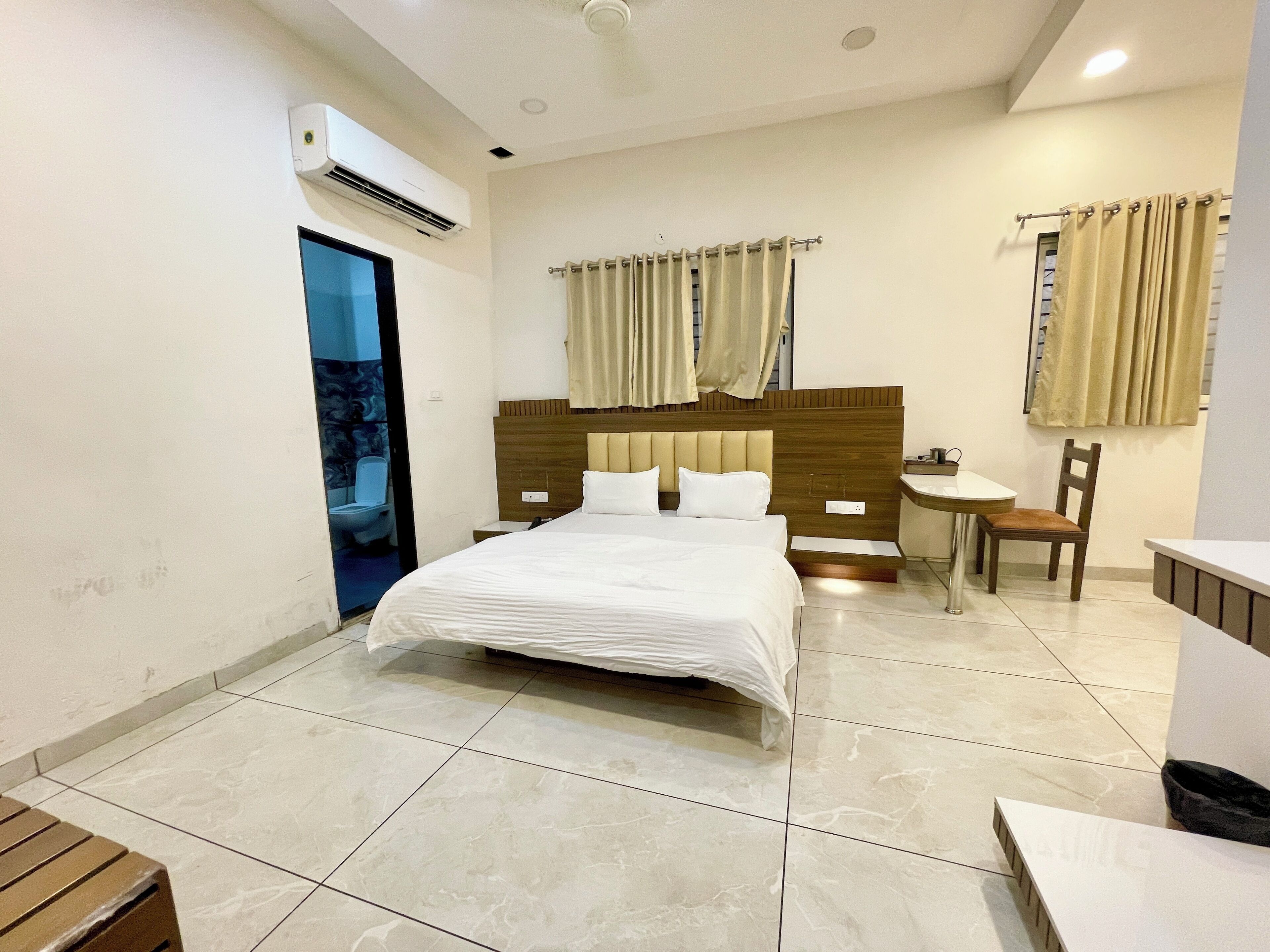 A hotel room with a large bed, a wooden headboard, a small table with a chair, a bathroom with a toilet and sink, and a window with curtains.