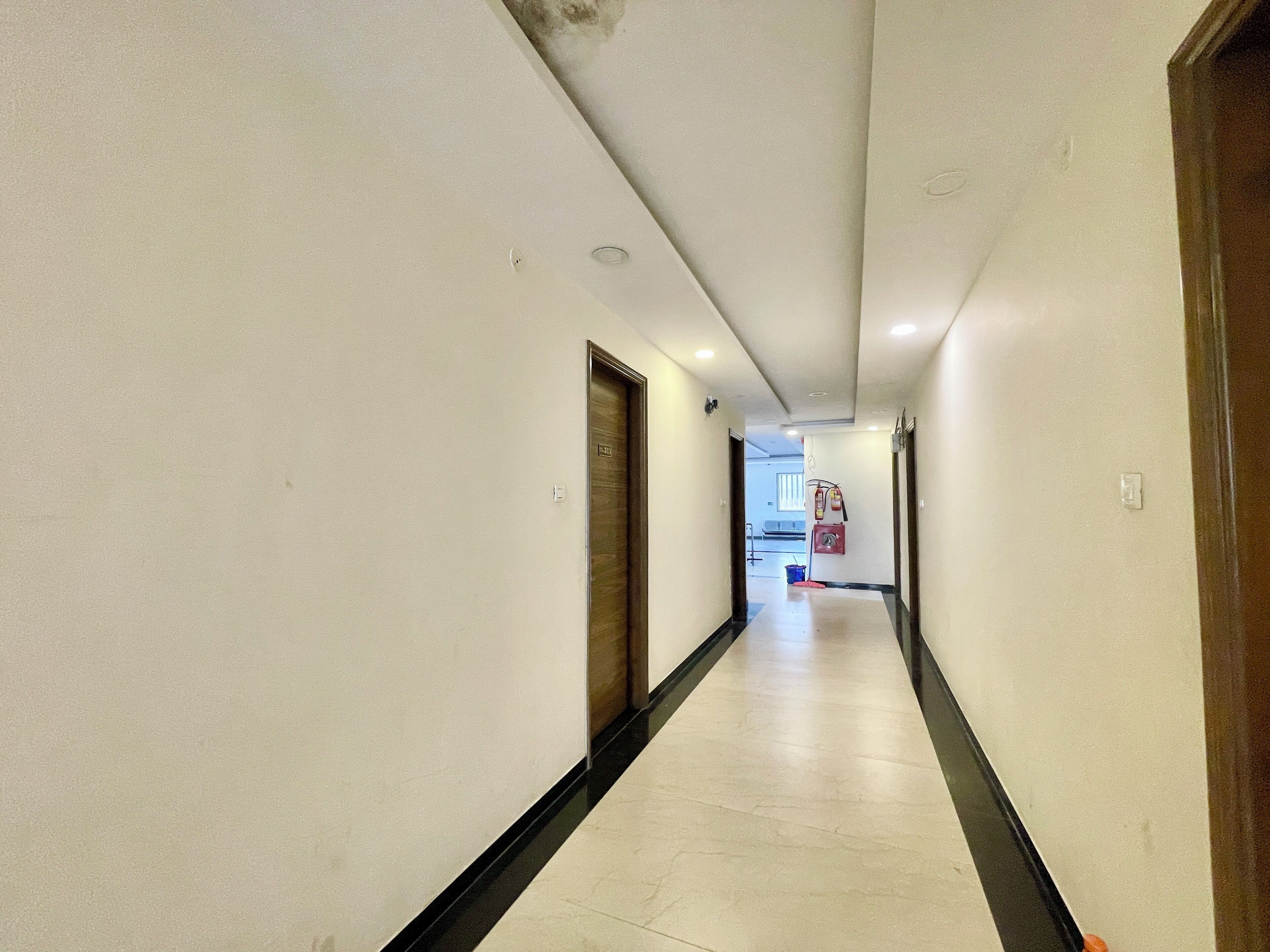 Ein Hotelgang mit weißen Wänden, einer schwarzen Fußleiste und einem gefliesten Boden. Auf der linken und rechten Seite befinden sich Türen, und eine Feuerlöscher ist an der Wand angebracht.