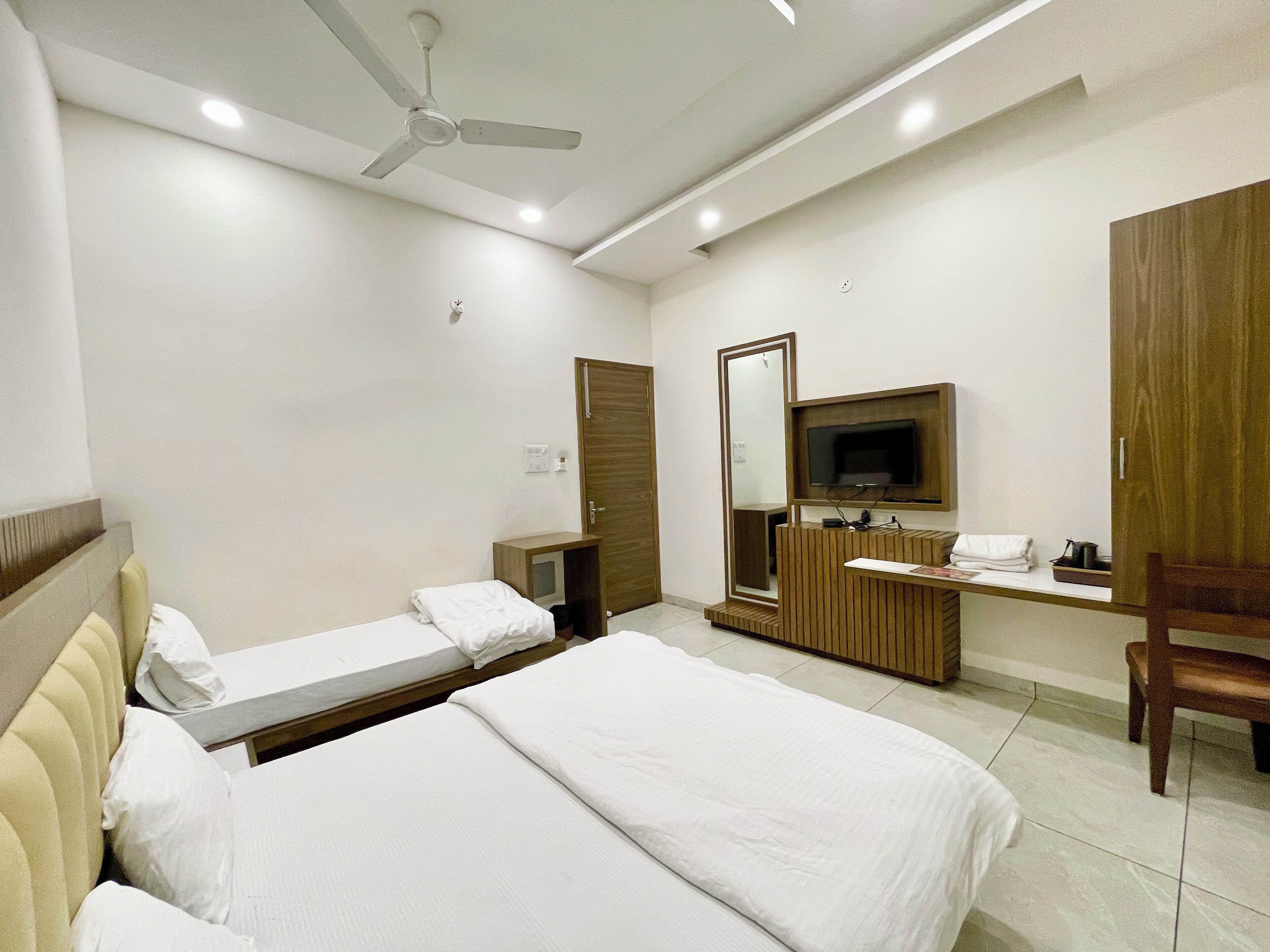 Une chambre d’hôtel avec deux lits, une télévision, un miroir, une armoire en bois et un ventilateur de plafond.