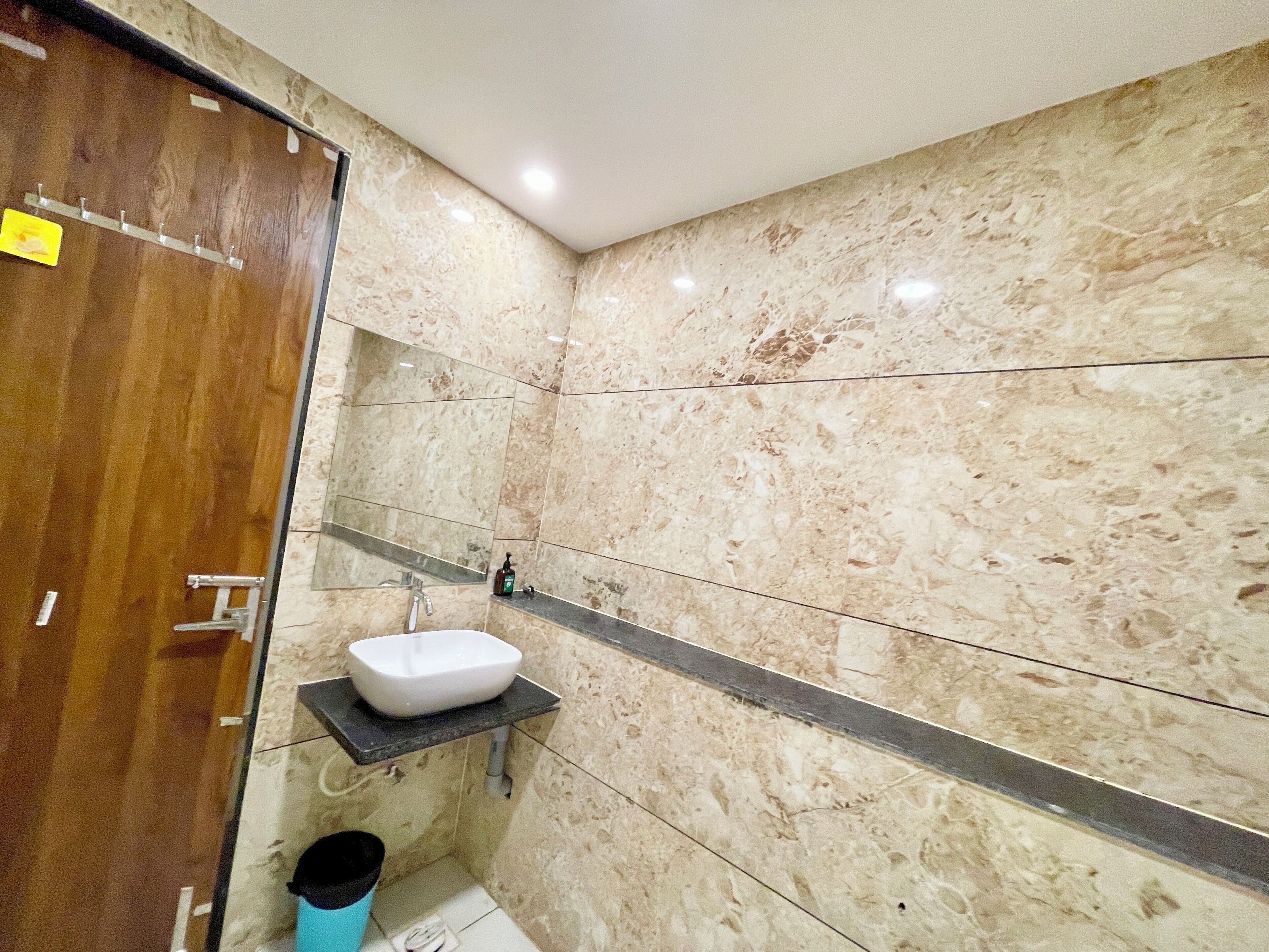 A bathroom with a wooden door, a white sink, a marble wall, and a small trash bin.