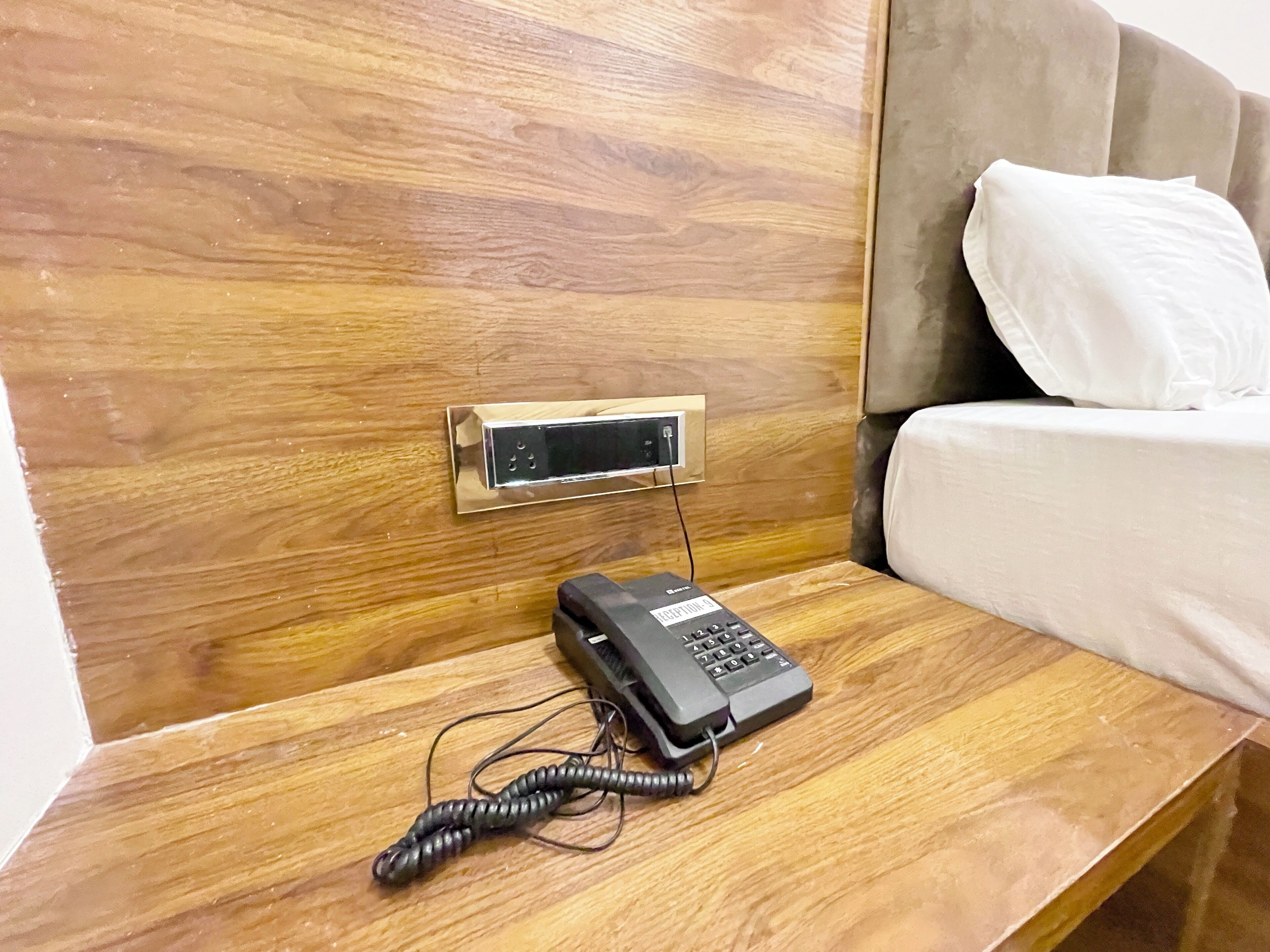 A wooden bedside table with a telephone, a power outlet, and a wall-mounted control panel.