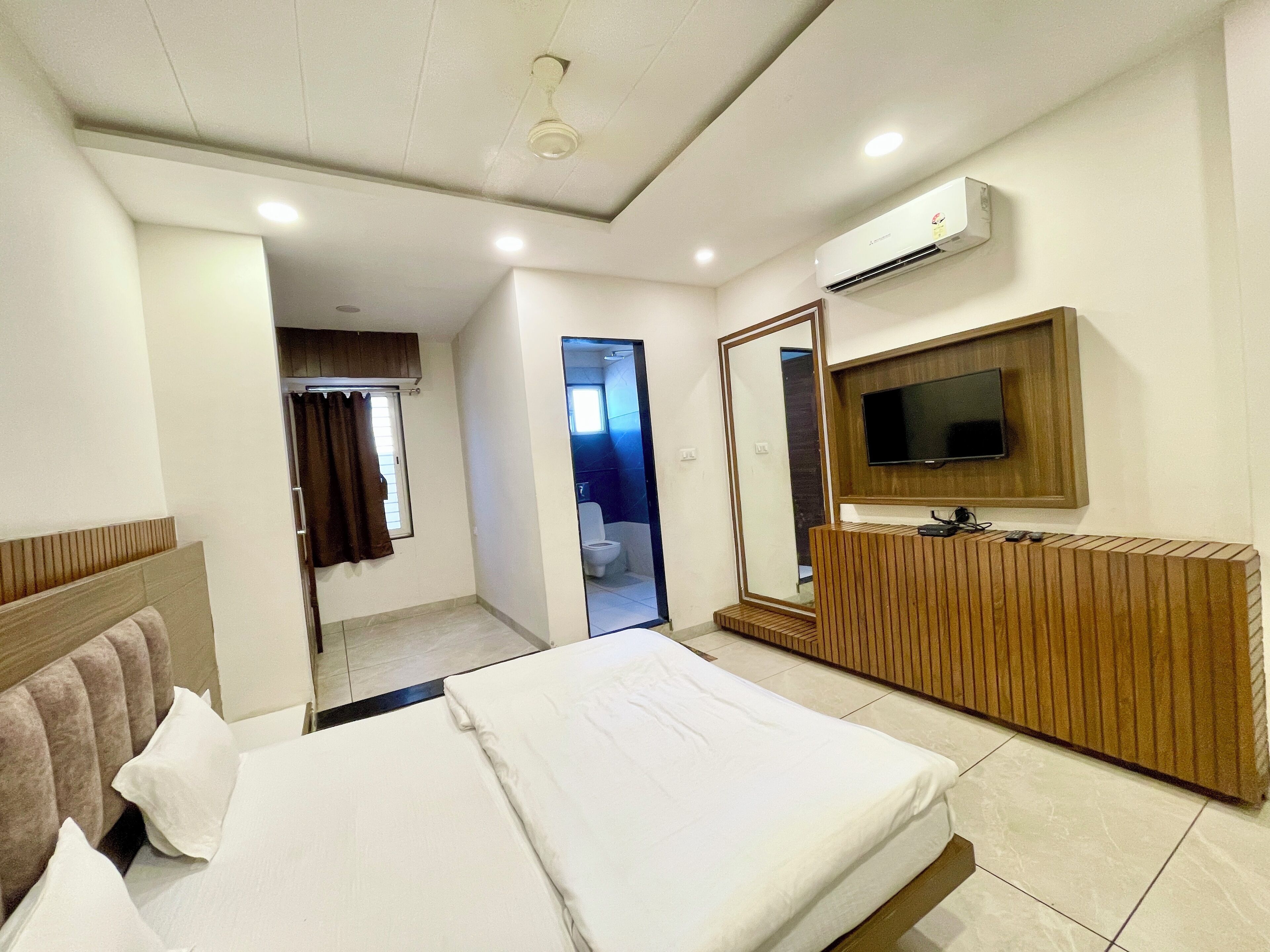 A hotel room with a bed, a TV mounted on the wall, a wooden dresser, and an air conditioning unit.