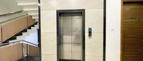 Un ascensor con panel de control digital, una puerta de madera con placa de número y una escalera con escalones color beige y barandilla de vidrio.