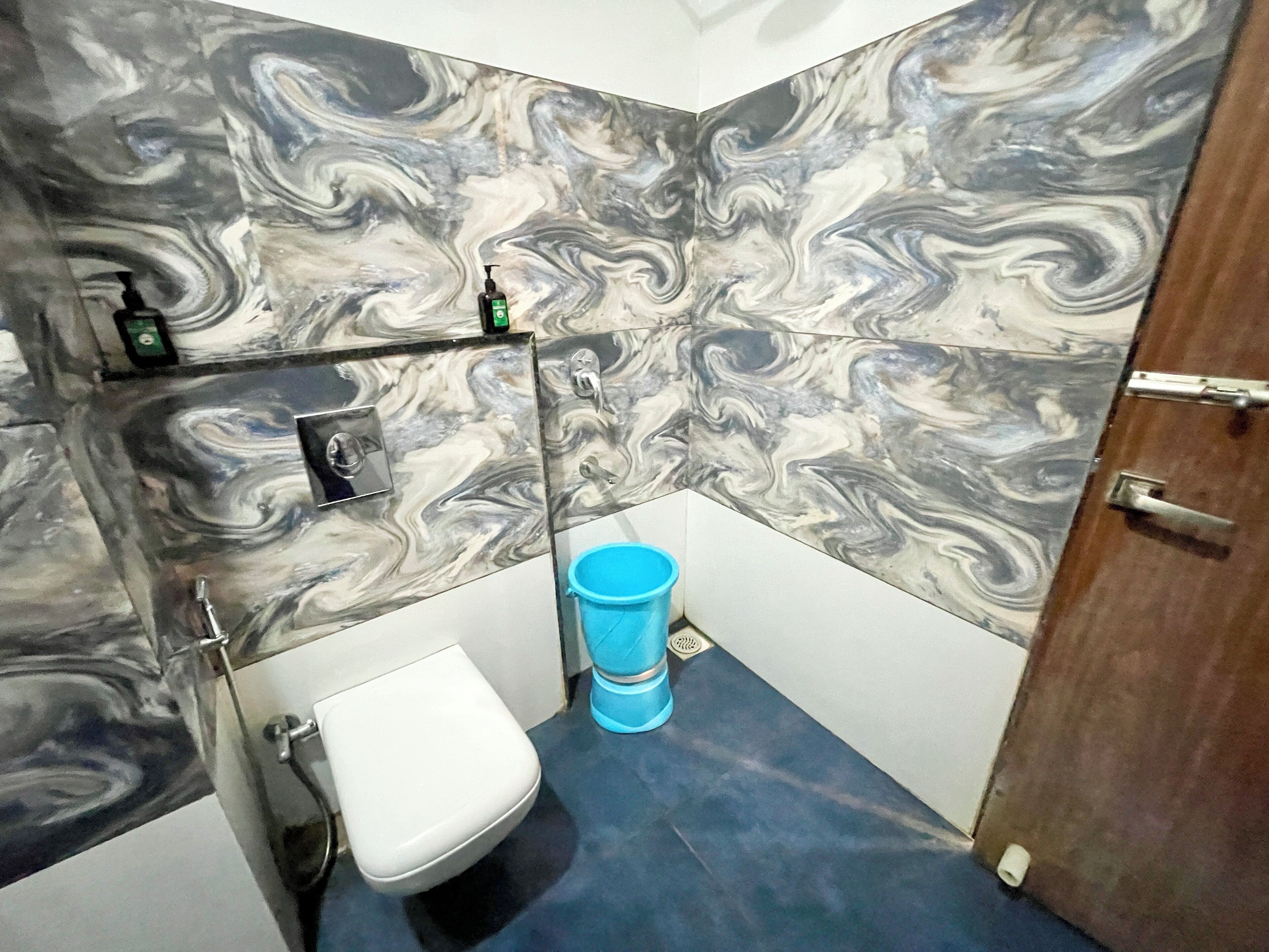 A bathroom with marble walls, a white toilet, a blue bucket, and a shelf with toiletries.