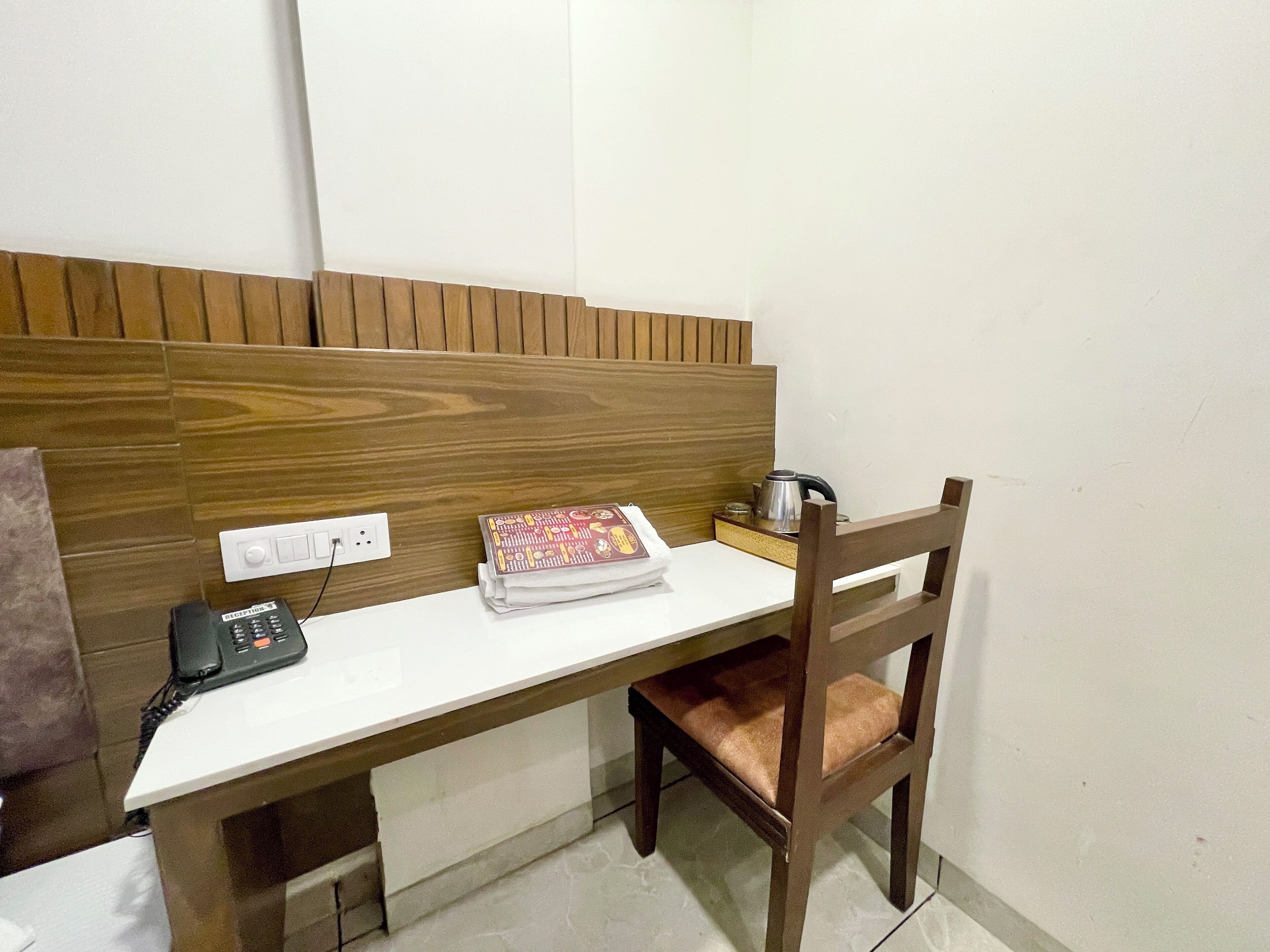 A hotel room with a wooden headboard, a desk with a telephone, a small table with a kettle, and a chair.