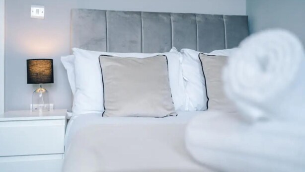 A neatly made bed with white and beige pillows, a grey headboard, and a bedside lamp with a black shade. - House 3 Bed Sleeps 6 -pets Ok -patio -free Parking (Manchester)