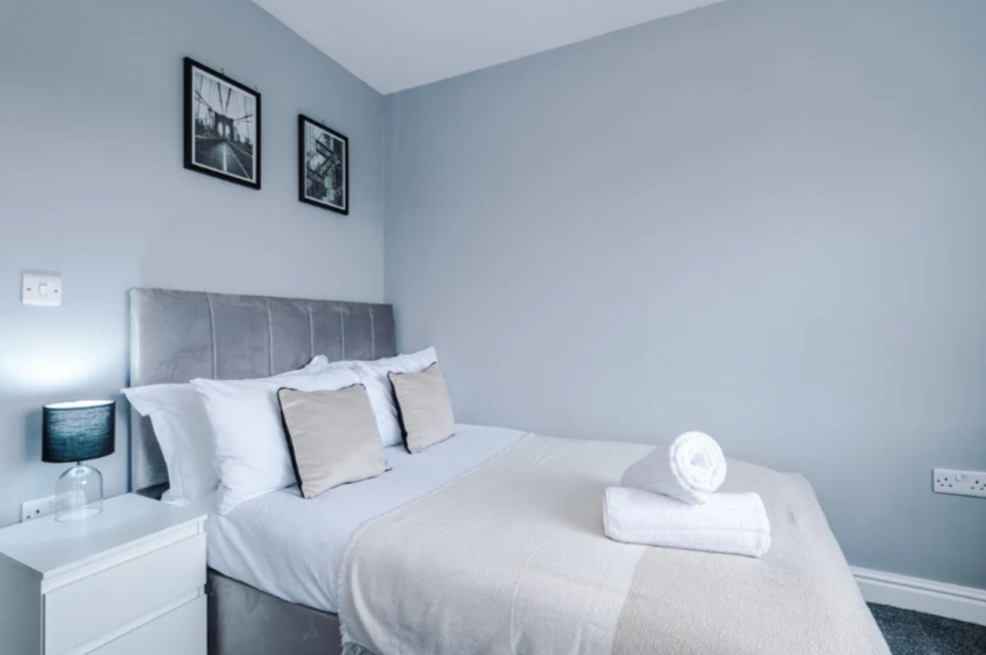 Quarto moderno com cabeceira cinza, roupa de cama branca e duas molduras na parede.
