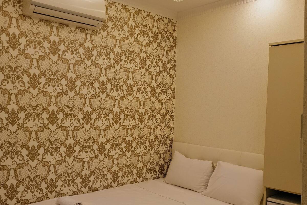 A hotel room with a bed, white linens, a decorative wall with a floral pattern, an air conditioning unit, and a small bedside table.