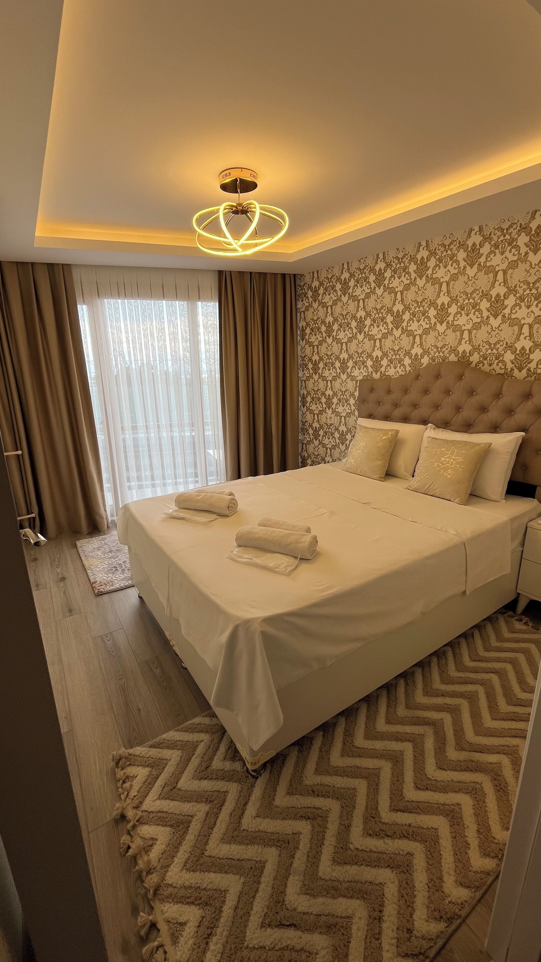 A neatly made bed with white linens and two rolled towels, a tufted headboard, a patterned wallpaper, and a chevron-patterned rug.