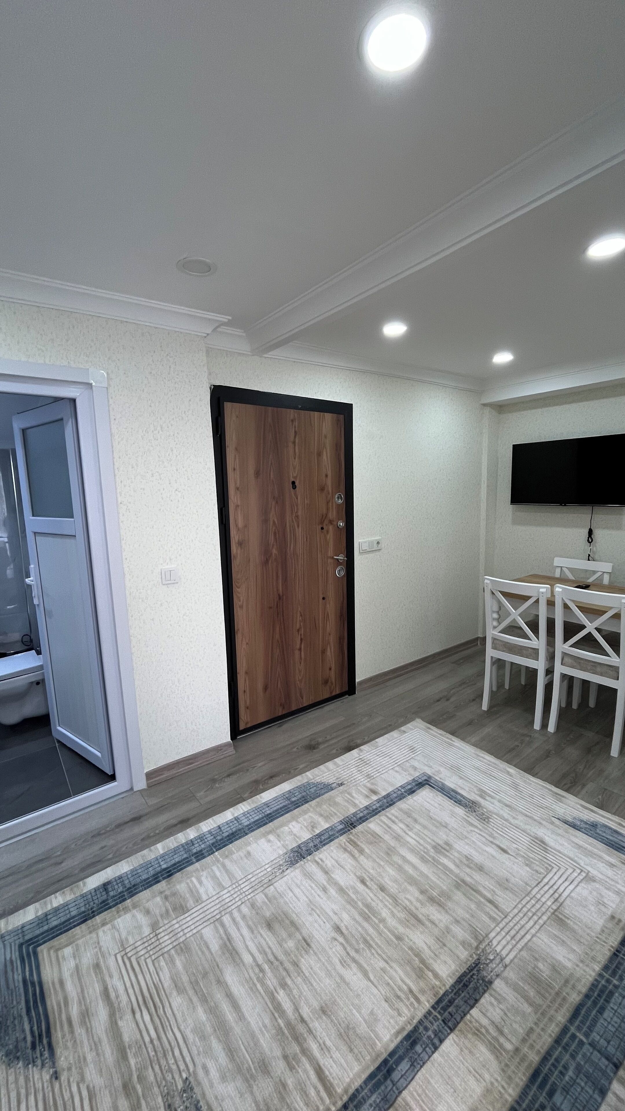 A room with a wooden door, white walls, a flat-screen TV mounted on the wall, a dining table with chairs, and a patterned rug on the floor.