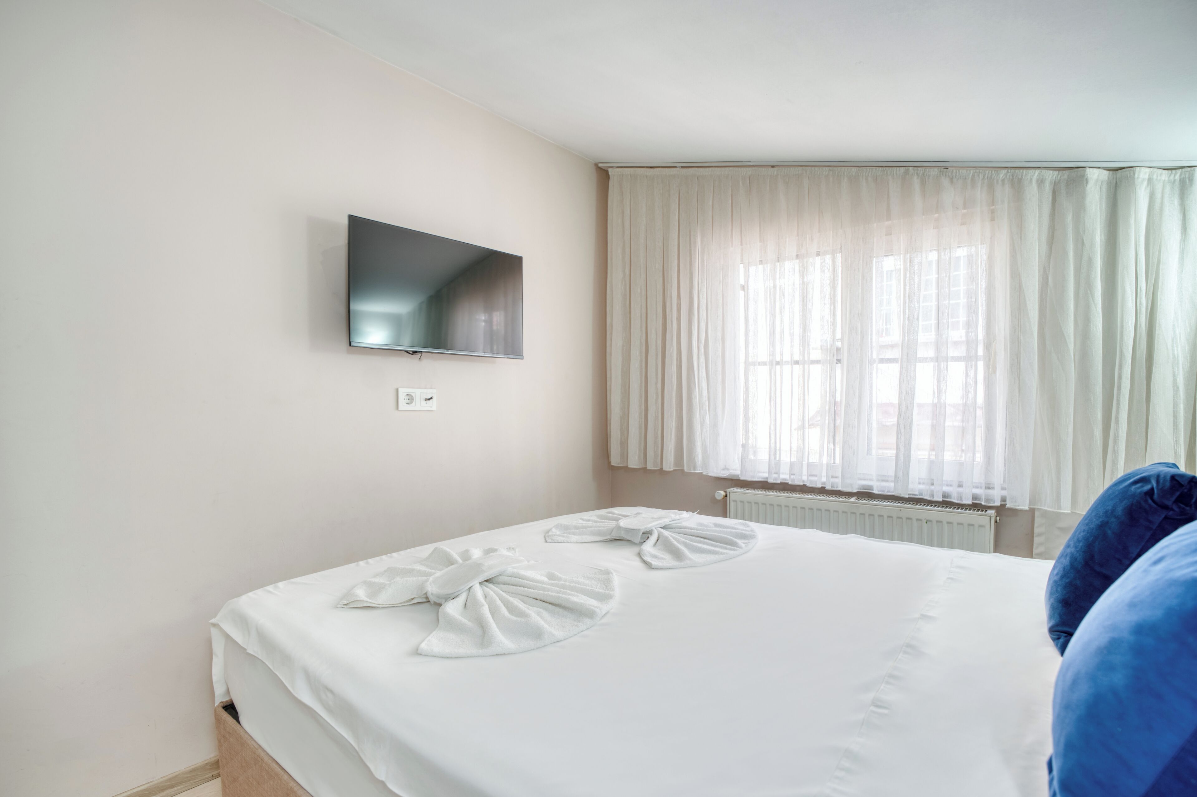 A hotel room with a bed, a flat-screen TV mounted on the wall, and a window with sheer curtains.