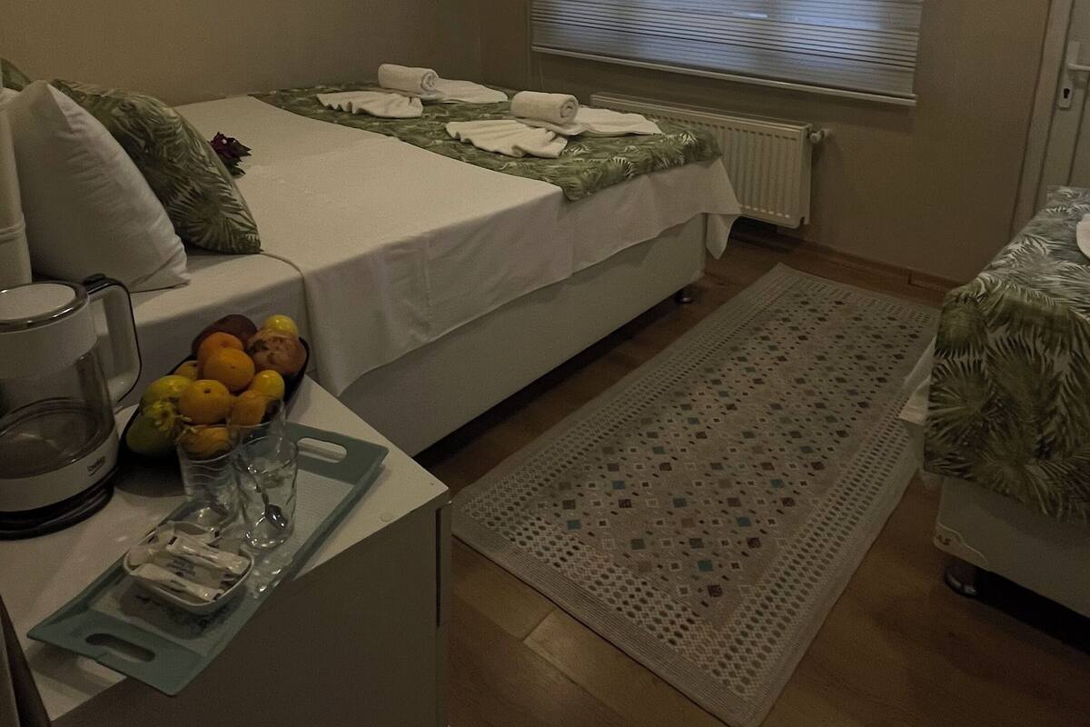 A hotel room with a bed, a bedside table with a coffee maker, a fruit bowl, and a window with blinds.