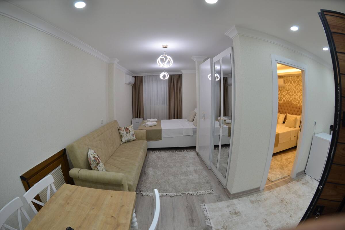 A modern living room with a sofa, a wooden table, and a glass partition leading to a bedroom with a bed and curtains.