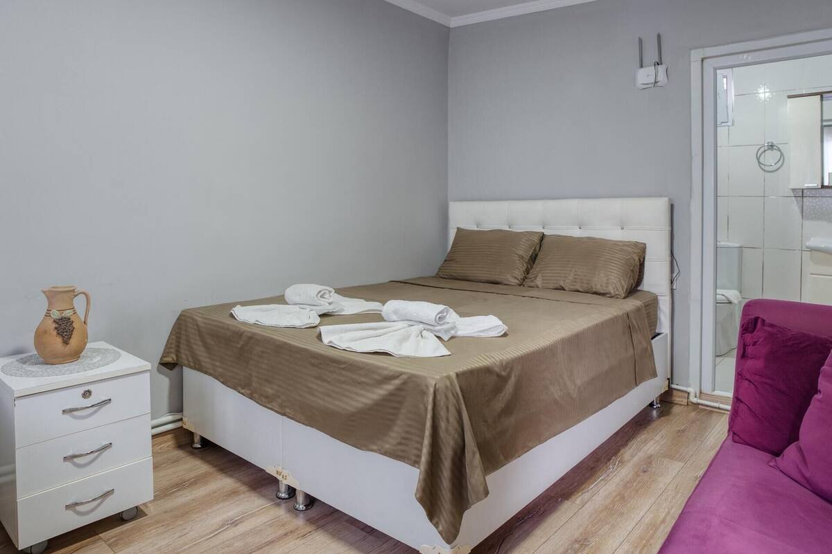 A bedroom with a bed, a bedside table with a vase, a drawer unit, and a purple chair.