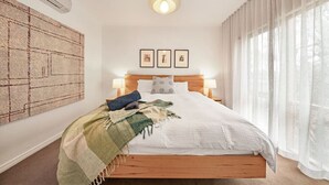 Un dormitorio con cama de madera, ropa de cama blanca, un tapiz grande tejido, tres cuadros enmarcados en la pared y cortinas traslúcidas en la ventana.