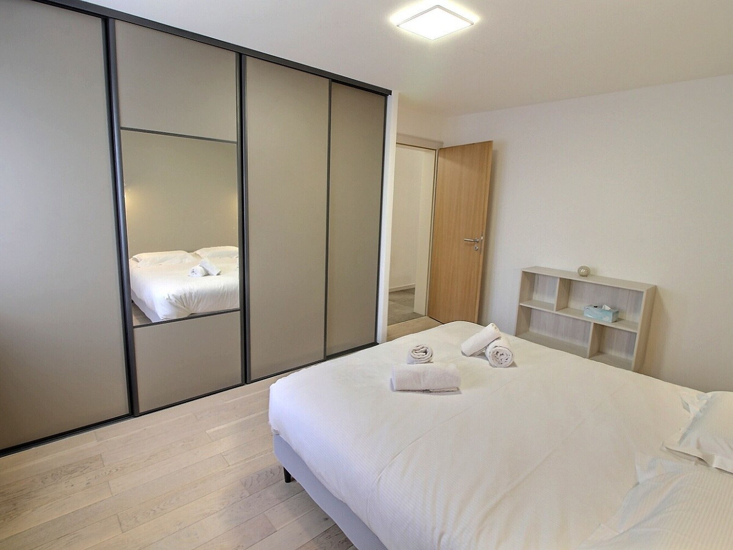 Une chambre d’hôtel moderne dotée d’un grand lit, d’une armoire avec des portes coulissantes, d’une table de chevet avec une lampe et d’une petite étagère contenant des articles de toilette.