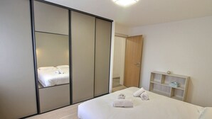 A modern hotel room with a large bed, a wardrobe with sliding doors, a bedside table with a lamp, and a small shelf with toiletries.