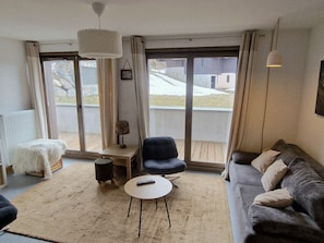 Un salon avec un canapé, un fauteuil, une petite table, une lampe et de grandes fenêtres.
