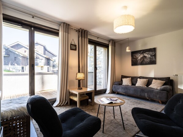 Un salón moderno con un sofá, dos sillones, una mesita y una lámpara. Hay un ventanal con cortinas, una pared de piedra y un cuadro en la pared.