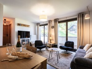 A modern living room with a wooden dining table, two black armchairs, a sofa, a television, and a large window with curtains.