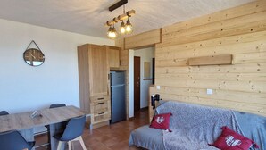 Un espacio habitable compacto con kitchenette, una zona de comedor con mesa y sillas de madera, y un sofá con manta y cojines.
