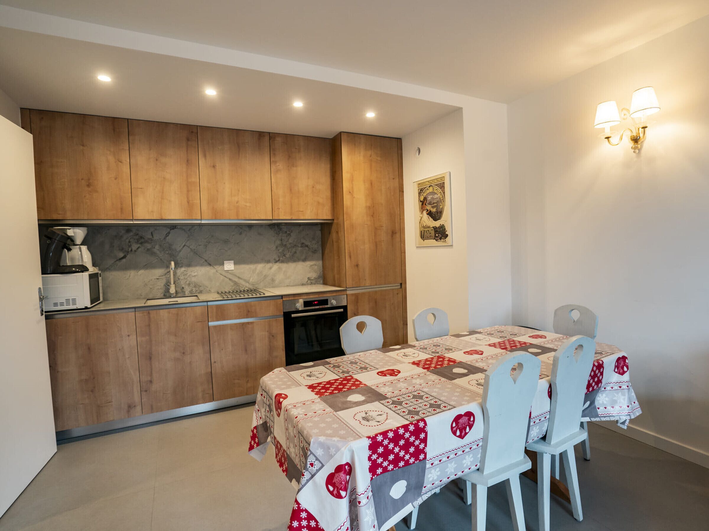 A compact kitchen with wooden cabinets, a microwave, and a coffee machine. There is a dining table with a quilted tablecloth and white chairs.