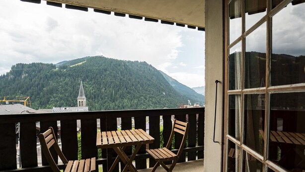 Un balcón con dos sillas y una mesa de madera, con vistas a un paisaje montañoso que incluye la aguja de una iglesia.