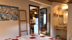 Un dormitorio con cama, colcha, mapa en la pared, baño con lavabo y espejo, y una puerta que da a otra habitación.