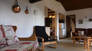 Una sala de estar acogedora con un sofá de madera, un sillón negro y una mesa de centro de madera. Se observa un área de cocina al fondo.
