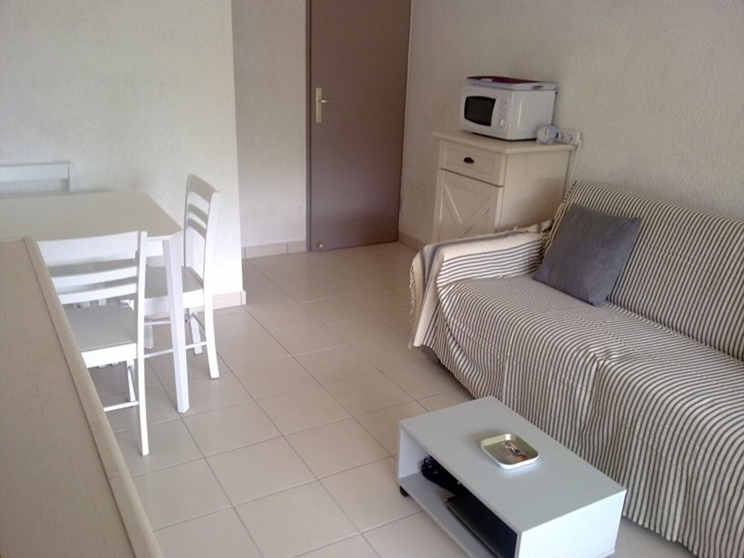 A small, clean room with a bed, a bedside table with a microwave, a chair, and a small table with a remote control.