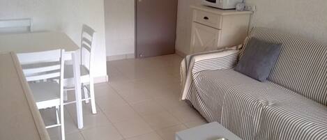 A small, clean room with a bed, a bedside table with a microwave, a chair, and a small table with a remote control.