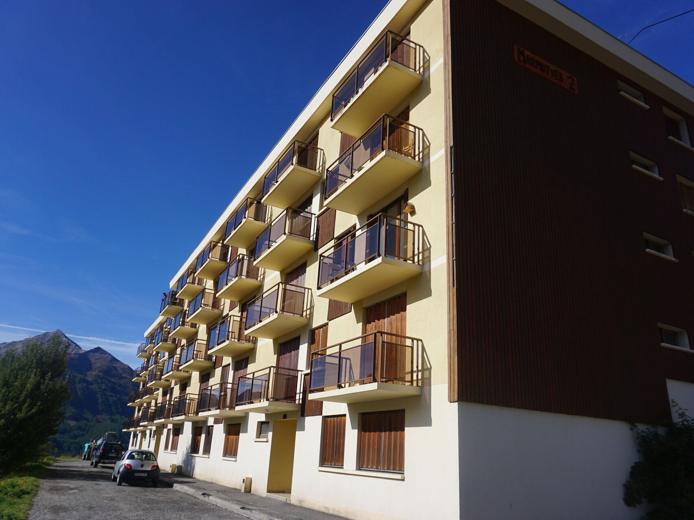 A multi-story hotel with balconies, a parking area, and a mountainous backdrop.