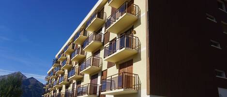 A multi-story hotel with balconies, a parking area, and a mountainous backdrop.