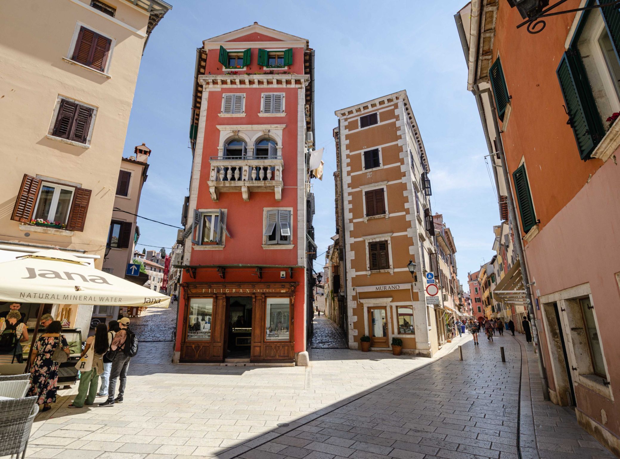 A narrow street with buildings featuring traditional architecture, including a prominent red building with a balcony and a sign that reads "MURANO".