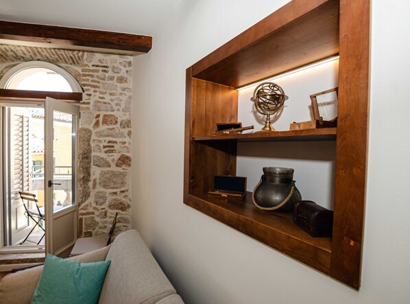 A cozy living area with a stone wall, wooden beams, a wooden shelf with decorative items, and a view through a glass door.