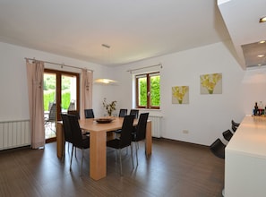 A modern dining area with a wooden table and black chairs, large windows with curtains, and a view of greenery outside.