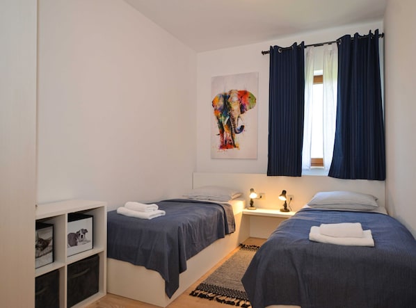 Un dormitorio con dos camas, una mesita de noche con lámparas, una ventana con cortinas, una obra de arte de un elefante y estantes de almacenamiento.