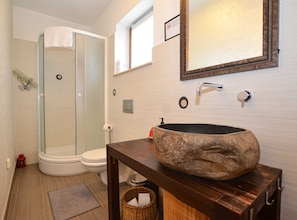A bathroom with a glass shower enclosure, a white toilet, a wooden vanity with a large stone sink, and a mirror above the sink.