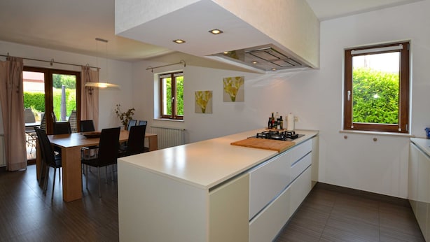 Une cuisine moderne dotée d’un îlot central, d’un coin repas avec une table et des chaises, et offrant une vue sur la verdure à travers les fenêtres.
