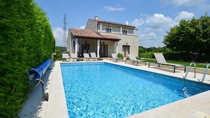 Une piscine entourée de chaises longues, un bâtiment en pierre surmonté d’un toit en tuiles et un ciel d’un bleu limpide.