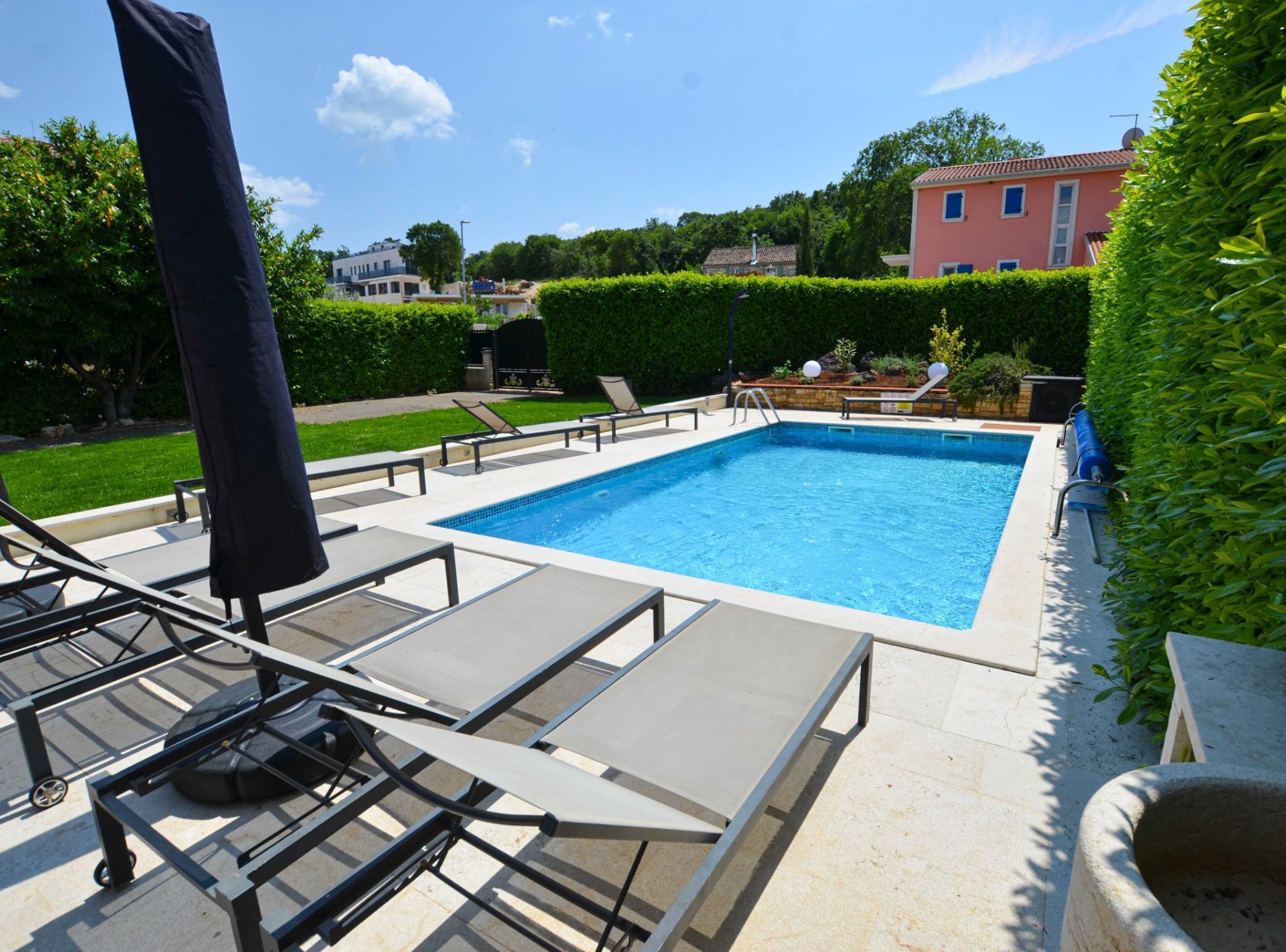 Ein rechteckiger Swimmingpool mit einer gefliesten Kante, umgeben von einer gepflasterten Liegefläche. Auf der Liegefläche befinden sich Liegestühle und ein Sonnenschirm.