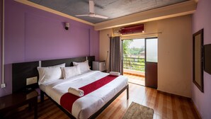 A modern bedroom with a large bed, a ceiling fan, a window with curtains, and a view of greenery outside. - Shree Rajeshwari Resort  (Morjim)