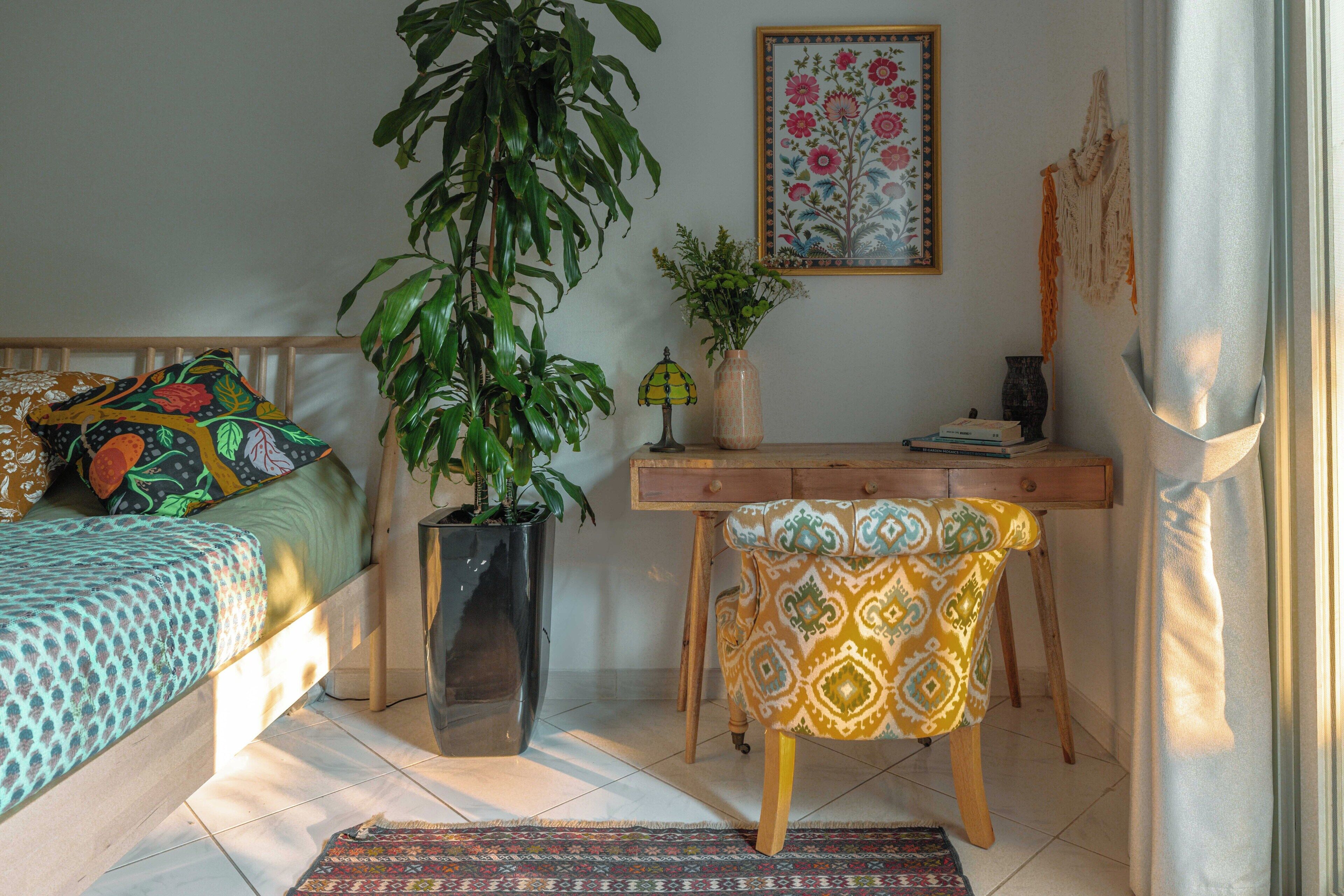 A cozy bedroom with a bed, a desk, a chair, a potted plant, and a window with curtains.