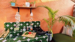 Un petit coin cuisine avec un mur recouvert de carreaux verts, une étagère en bois ornée de plantes en pot, un luminaire suspendu et un banc vert avec un coussin.