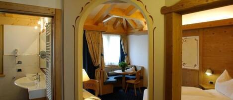 Habitación de hotel con techo de madera, un pequeño comedor con mesa y sillas, una ventana con cortinas y un baño con lavamanos y espejo.