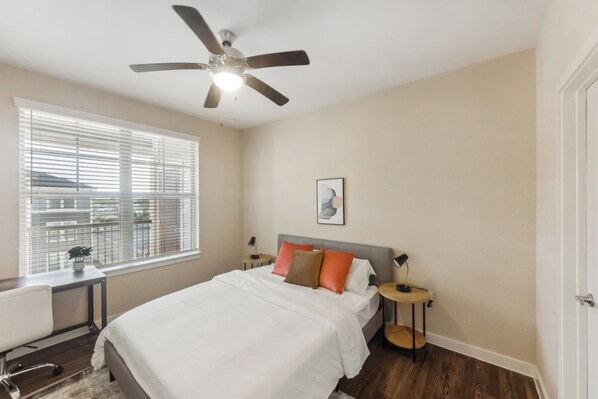 A bedroom with a bed, a desk, a chair, a ceiling fan, a window with blinds, and a nightstand with a lamp. - Charming & Modern 1Bd/1Bth I Properties by Preston (Houston)