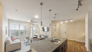 A modern kitchen with granite countertops, stainless steel sinks, and a dishwasher. There is a dining area with a table and chairs, and a living room with a sofa and a TV. - Cullen 5312 I New Construction 2Bd Apt I Gated (Houston)