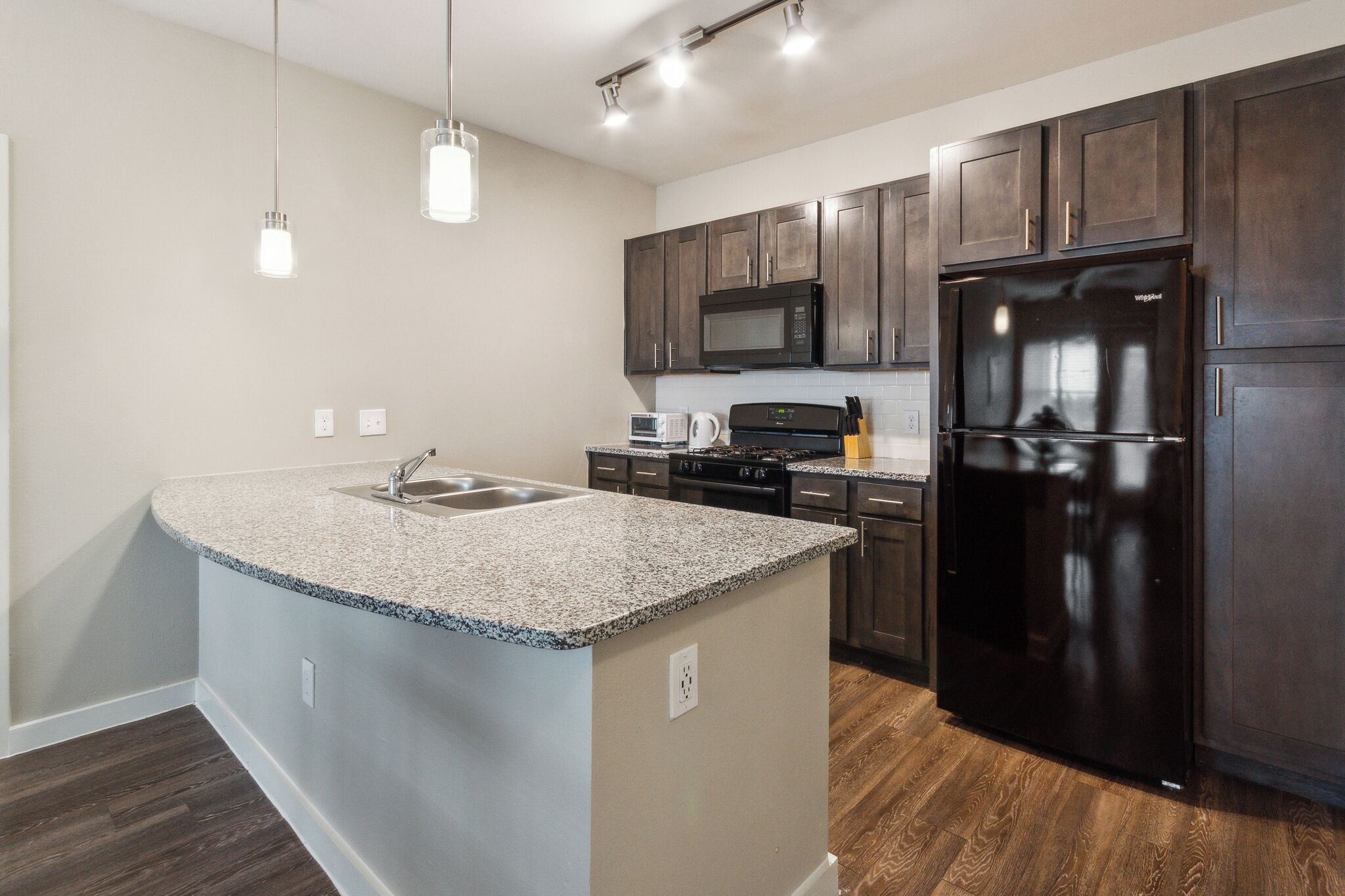 A modern kitchen with dark wood cabinets, a granite countertop, stainless steel appliances, and pendant lighting.